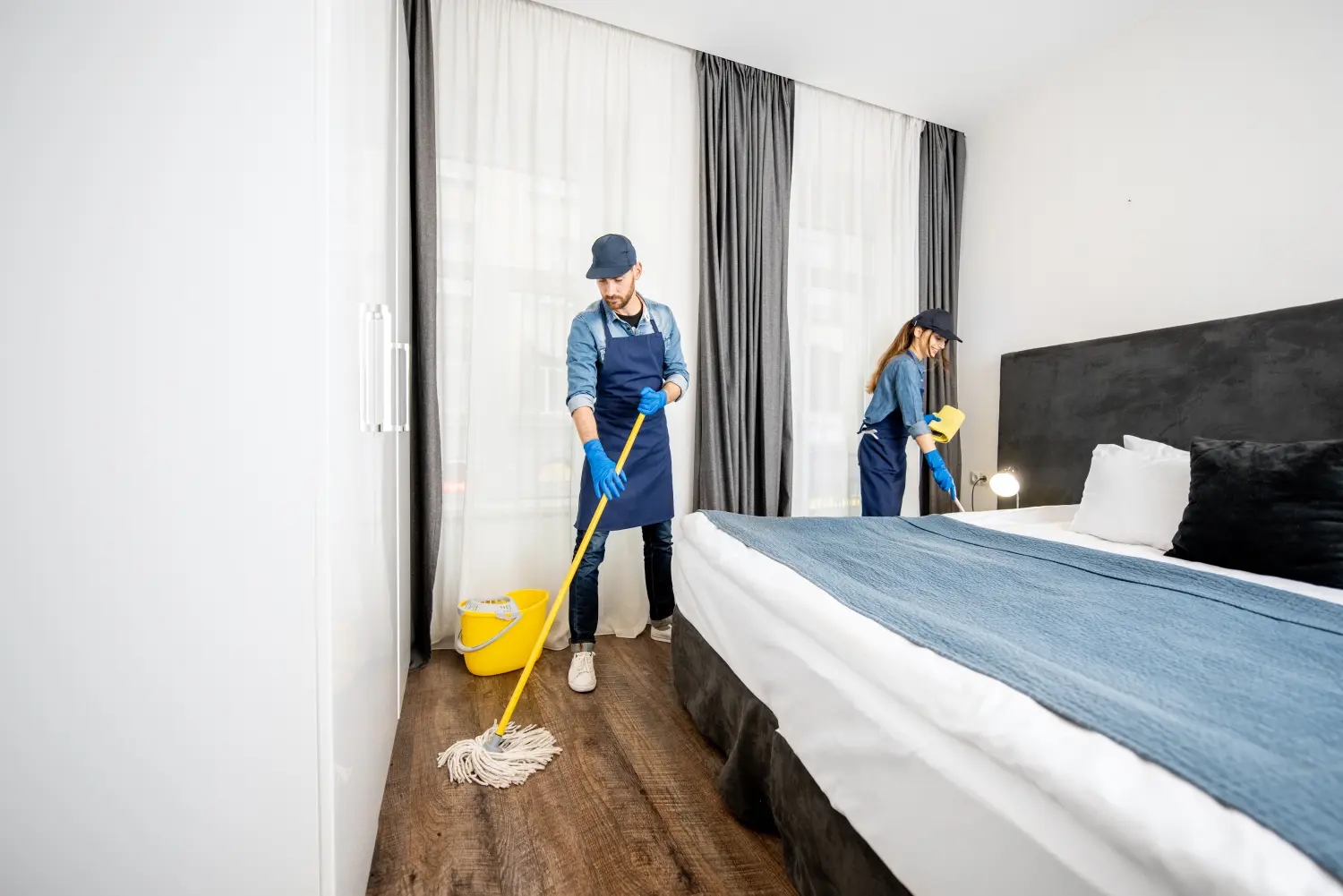 Professionelle Reinigungskräfte in Uniform waschen den Boden und wischen Staub von den Möbeln im Hotelzimmer oder Schlafzimmer.